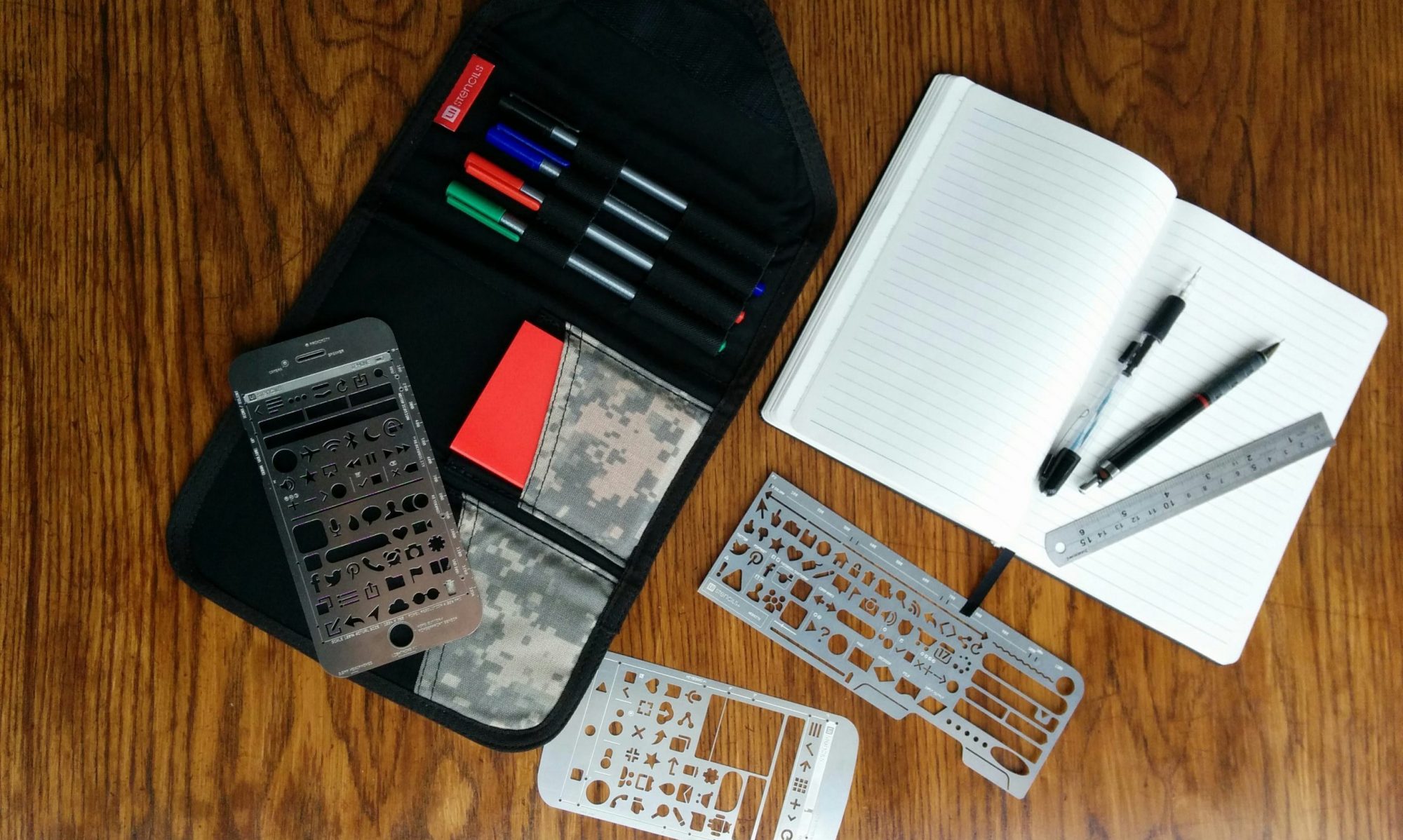 A selection of UX stencils, markers, pencils, and a notebook on a wooden desk surface