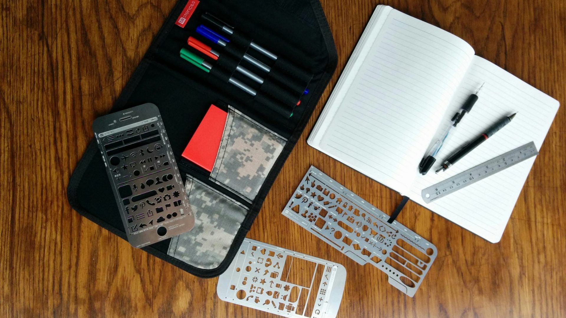 A selection of UX stencils, markers, pencils, and a notebook on a wooden desk surface